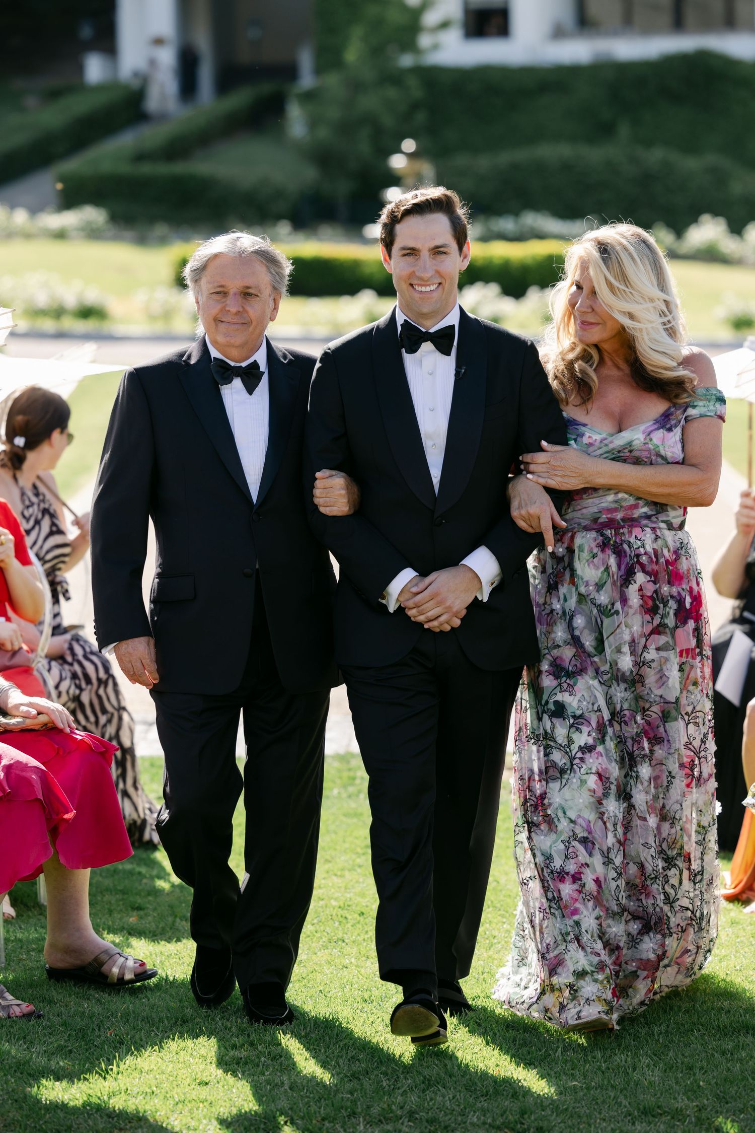 Wedding party members in formal attire walk and laugh together on a sunny lawn.