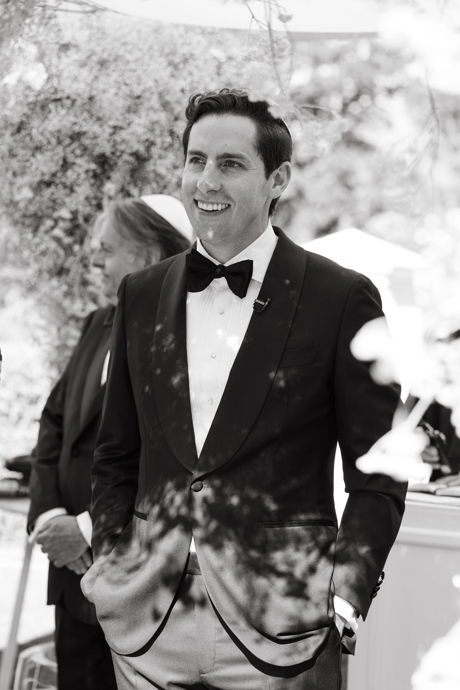 Black and white photo of a person in a tuxedo and bow tie smiling at an outdoor event.