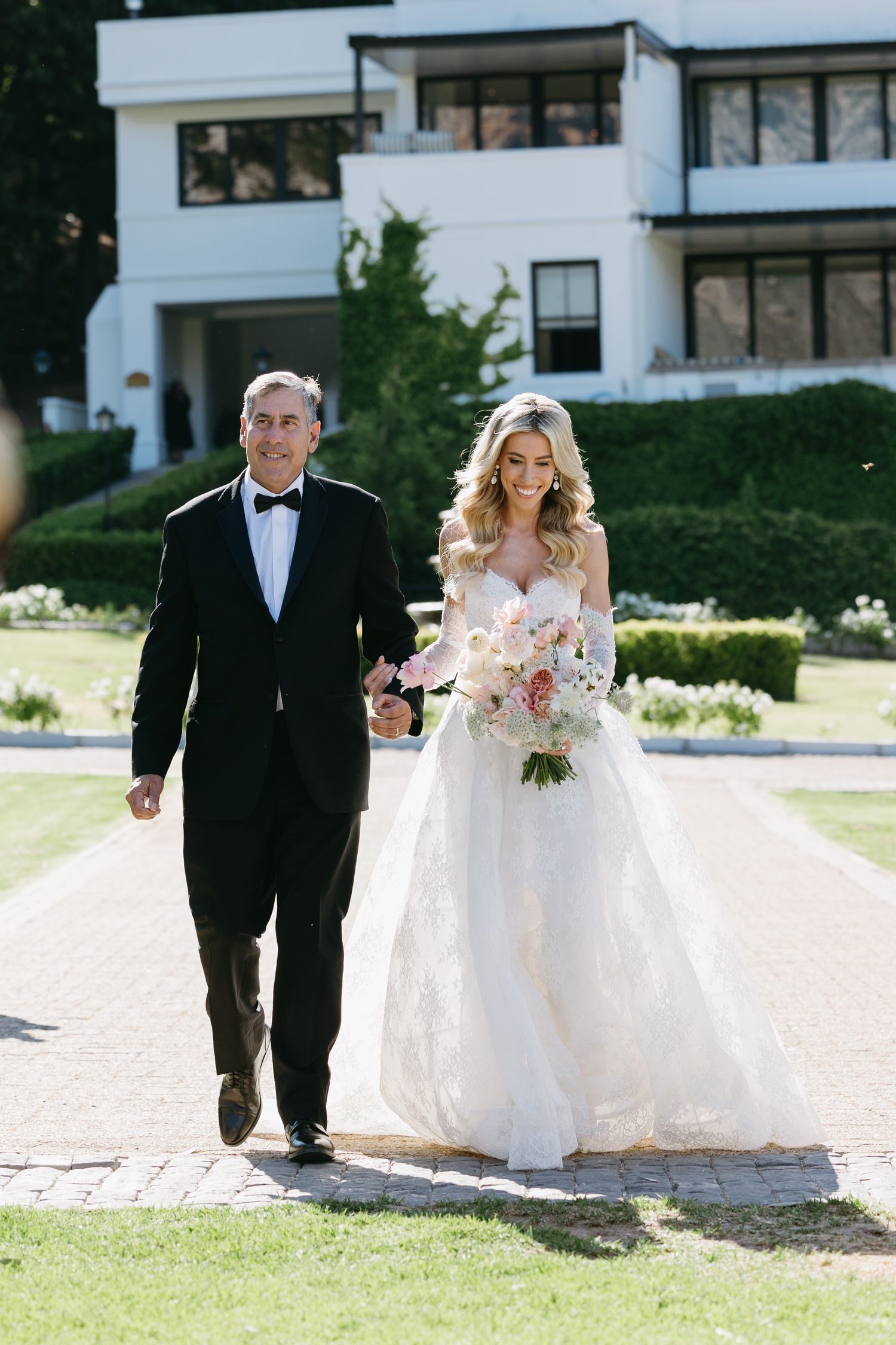 A wedding processional on a sunny day with a formal black tuxedo and flowing white bridal gown outside an elegant mansion.