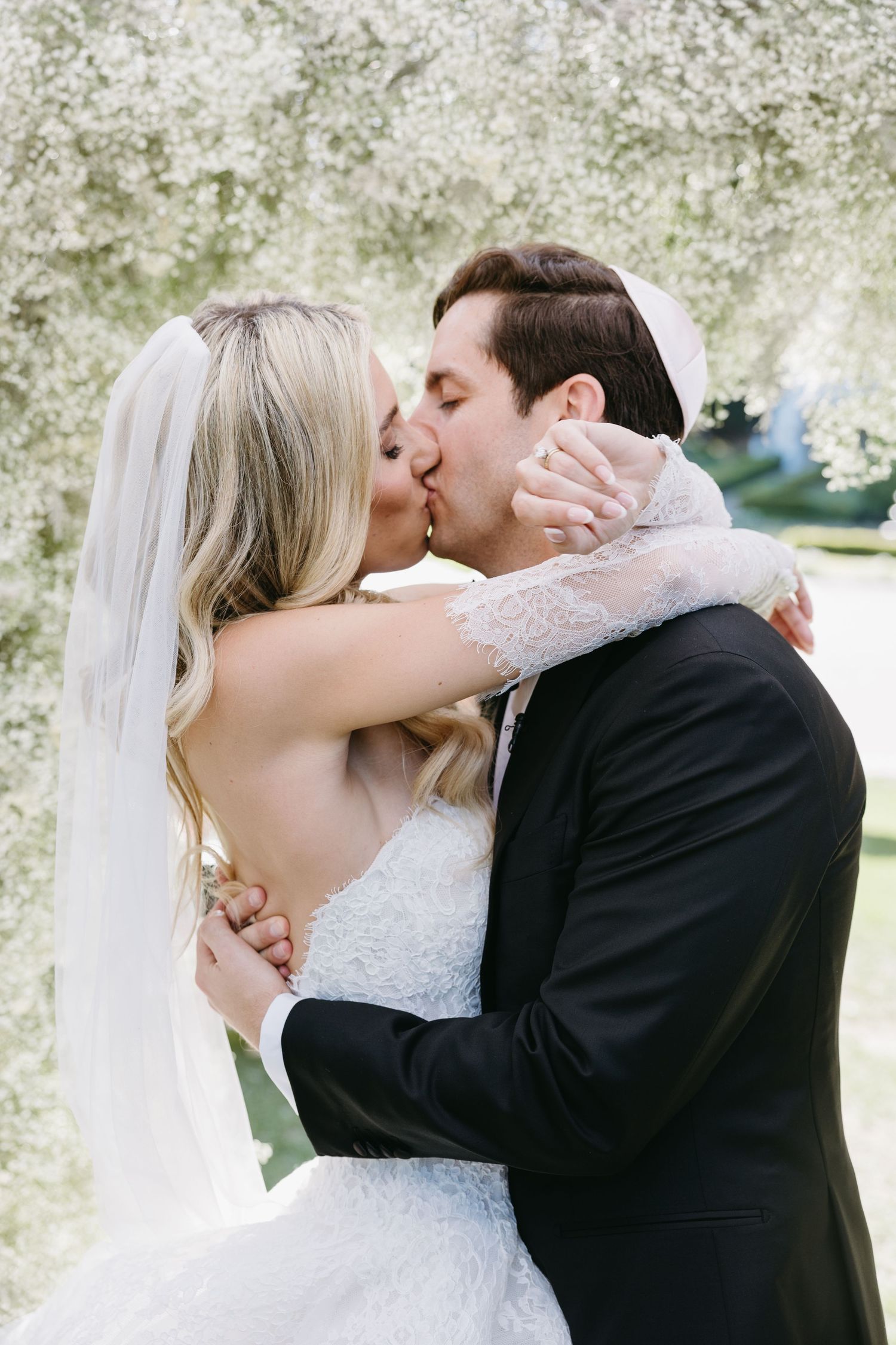 Newlyweds share their first kiss as husband and wife under a canopy of white blossoms.