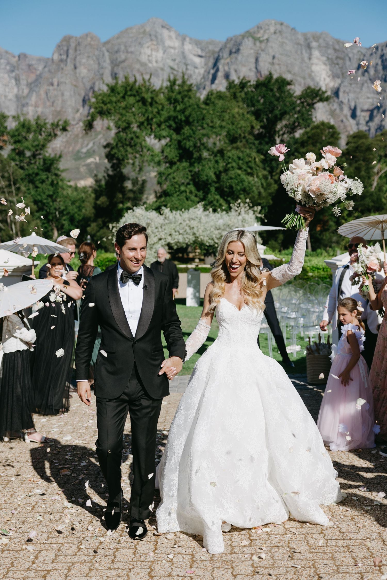 A joyful wedding celebration unfolds against a backdrop of dramatic mountain cliffs on a sunny day.