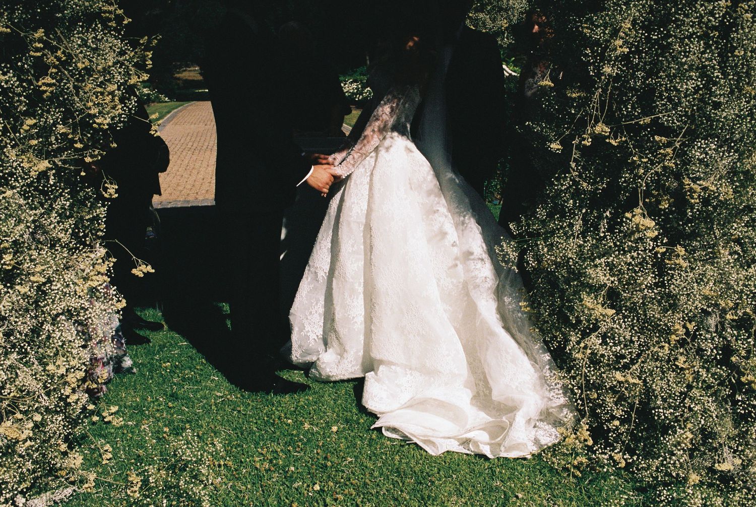 Wedding guests gather on a grassy lawn under flowering trees during an outdoor ceremony.