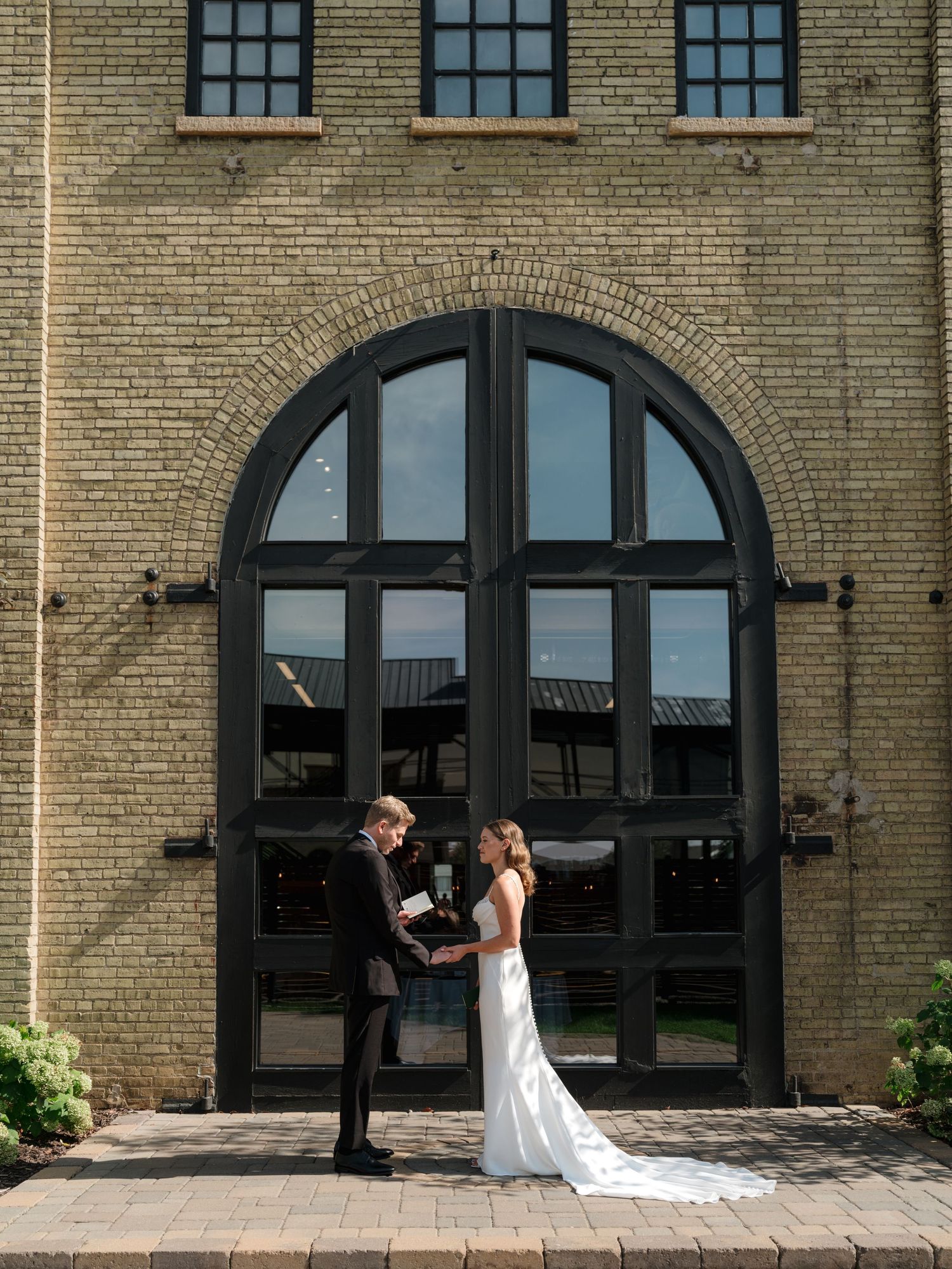 Couple shares a romantic moment in front of a historic brick building with arched windows and doorways.