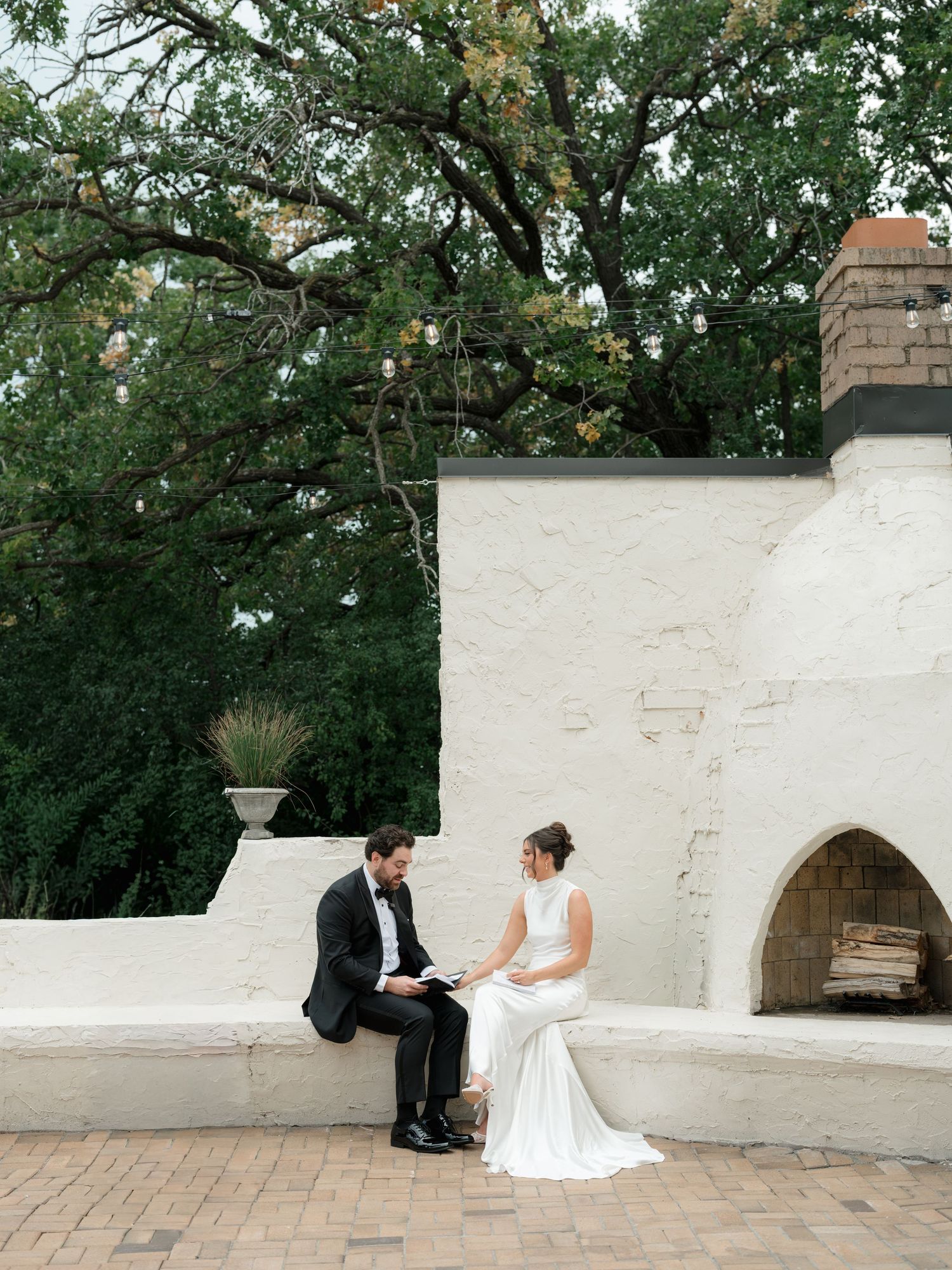 Couple in wedding attire sits together on white stone steps in a lush garden setting.