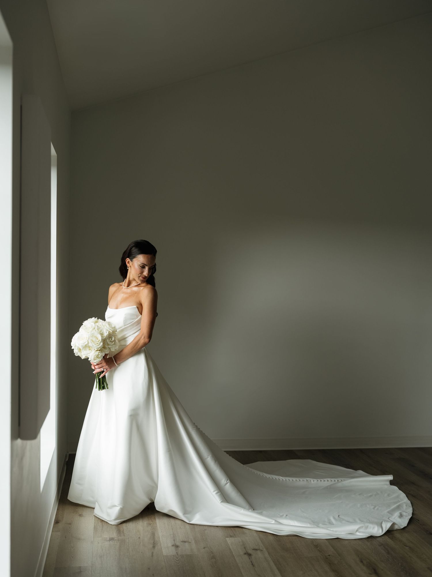 A bride in a flowing white wedding gown poses elegantly against a minimalist wall with dramatic lighting.