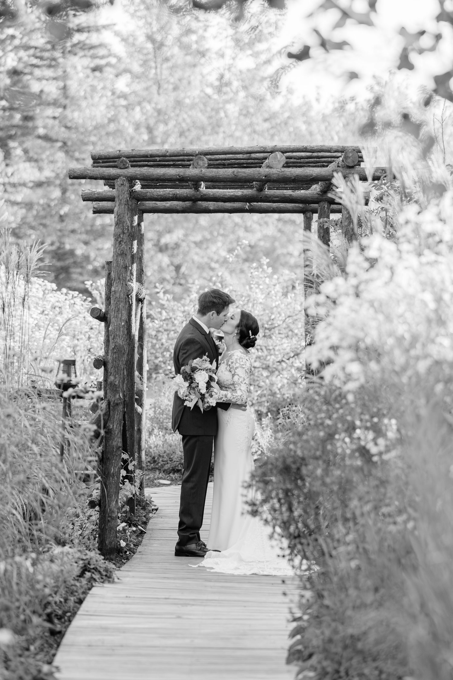 A romantic black and white wedding photo showing a couple embracing under a rustic wooden arbor on a garden pathway.