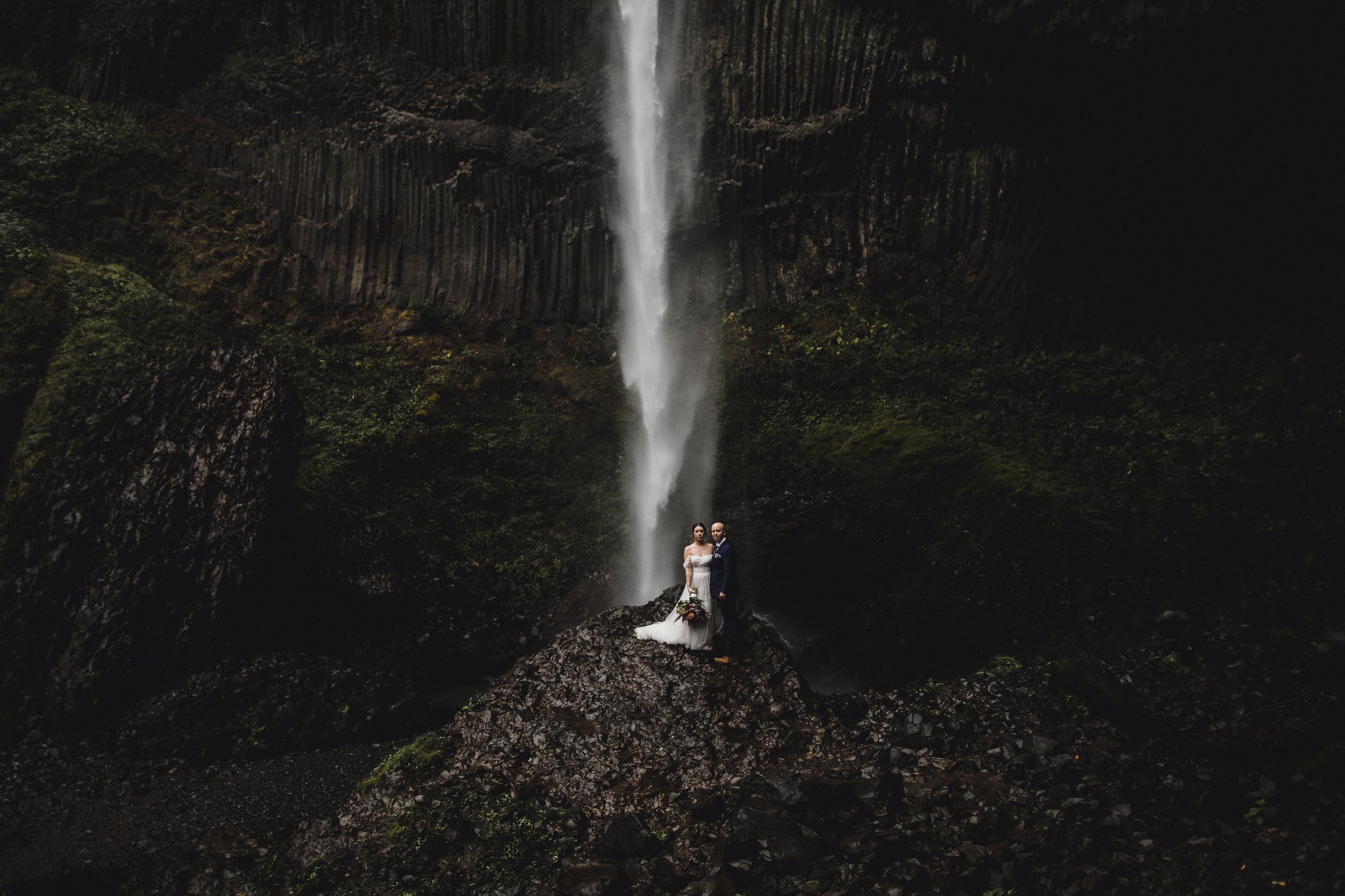 A dramatic waterfall cascades down a dark rocky cliff face against a moody forest backdrop.