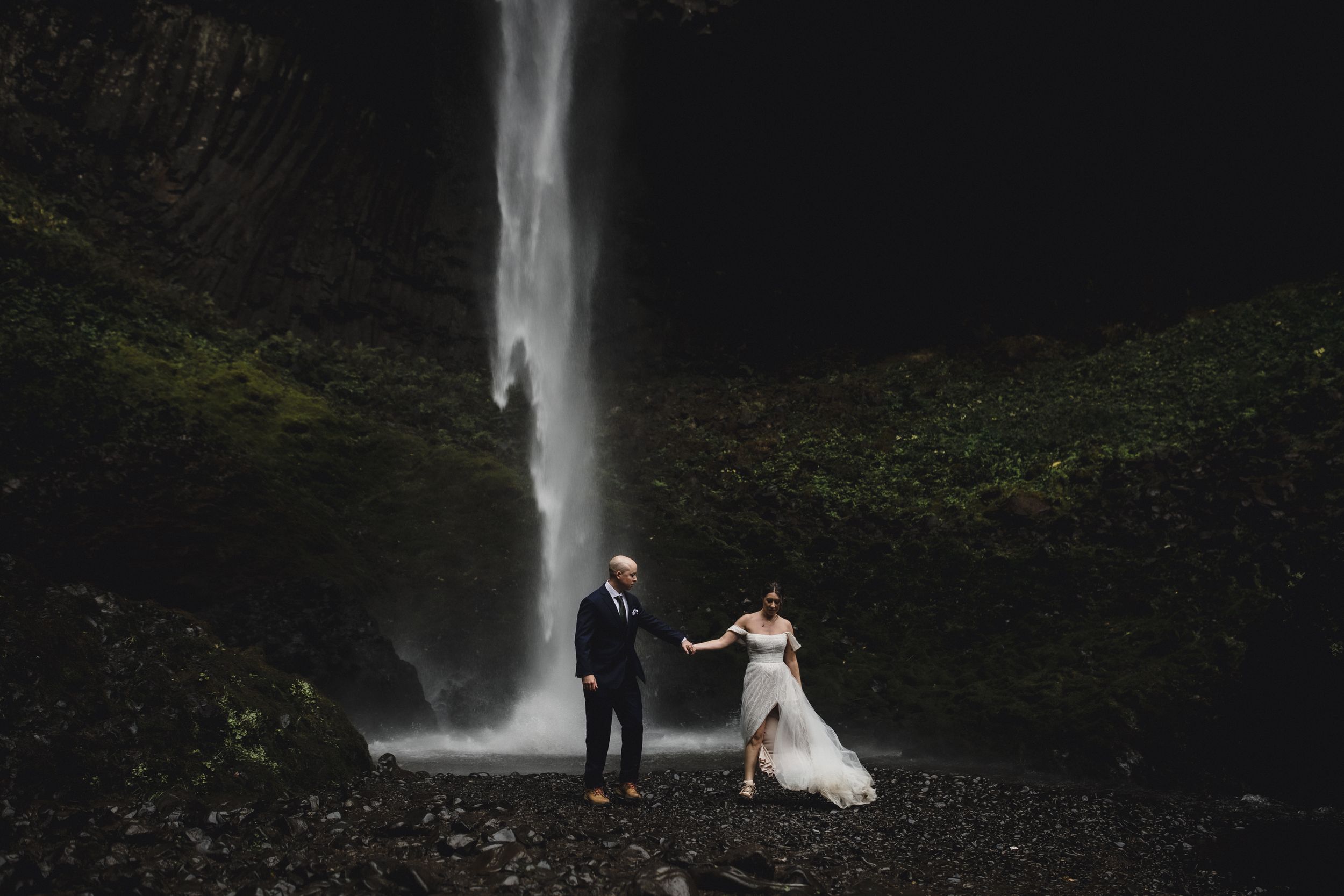 A couple poses in wedding attire near a majestic waterfall surrounded by lush greenery.