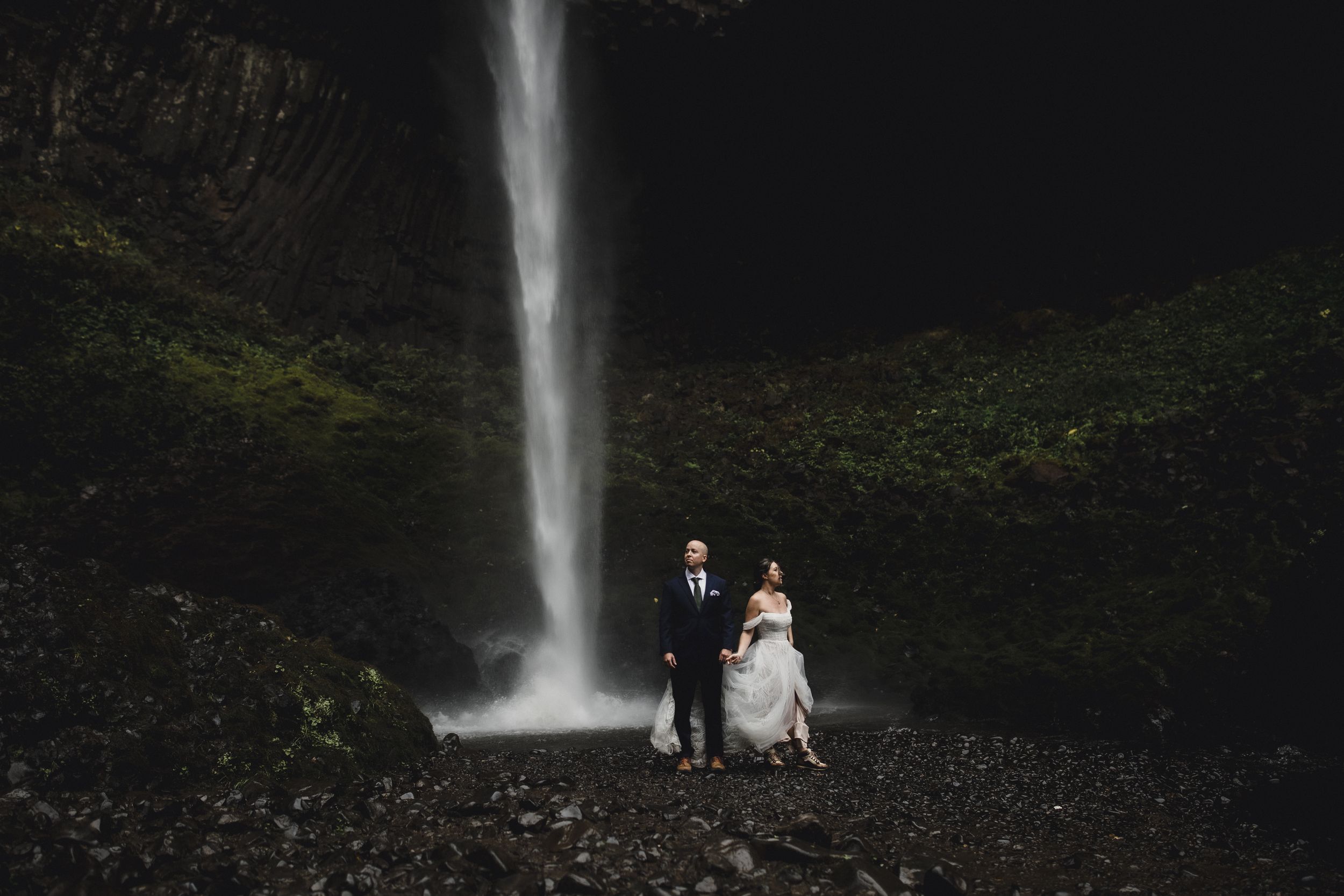 Two figures stand before a towering waterfall in formal attire amid a dramatic natural landscape.