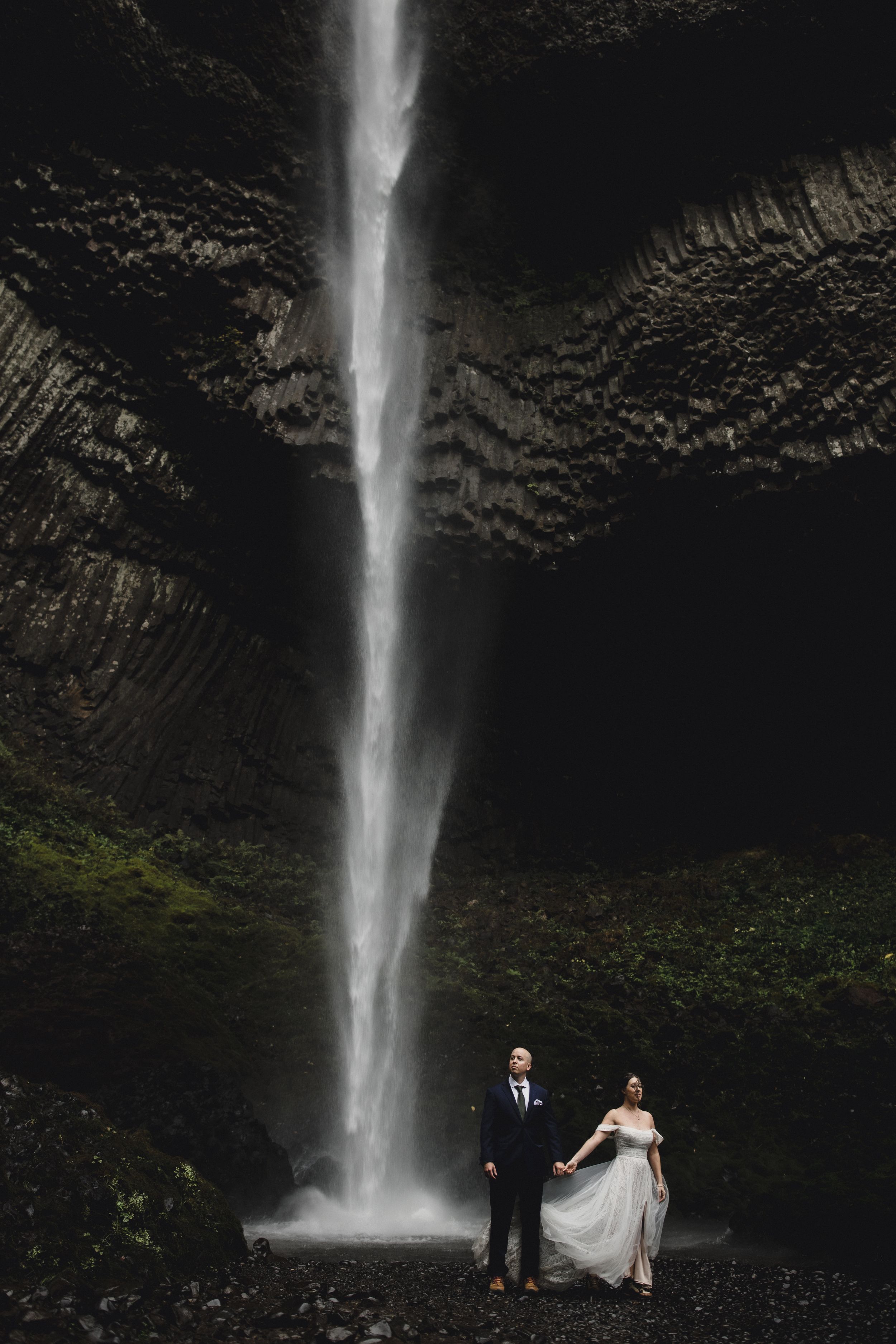 A dramatic waterfall cascades down black rock cliffs in a moody landscape with two people standing at its base.