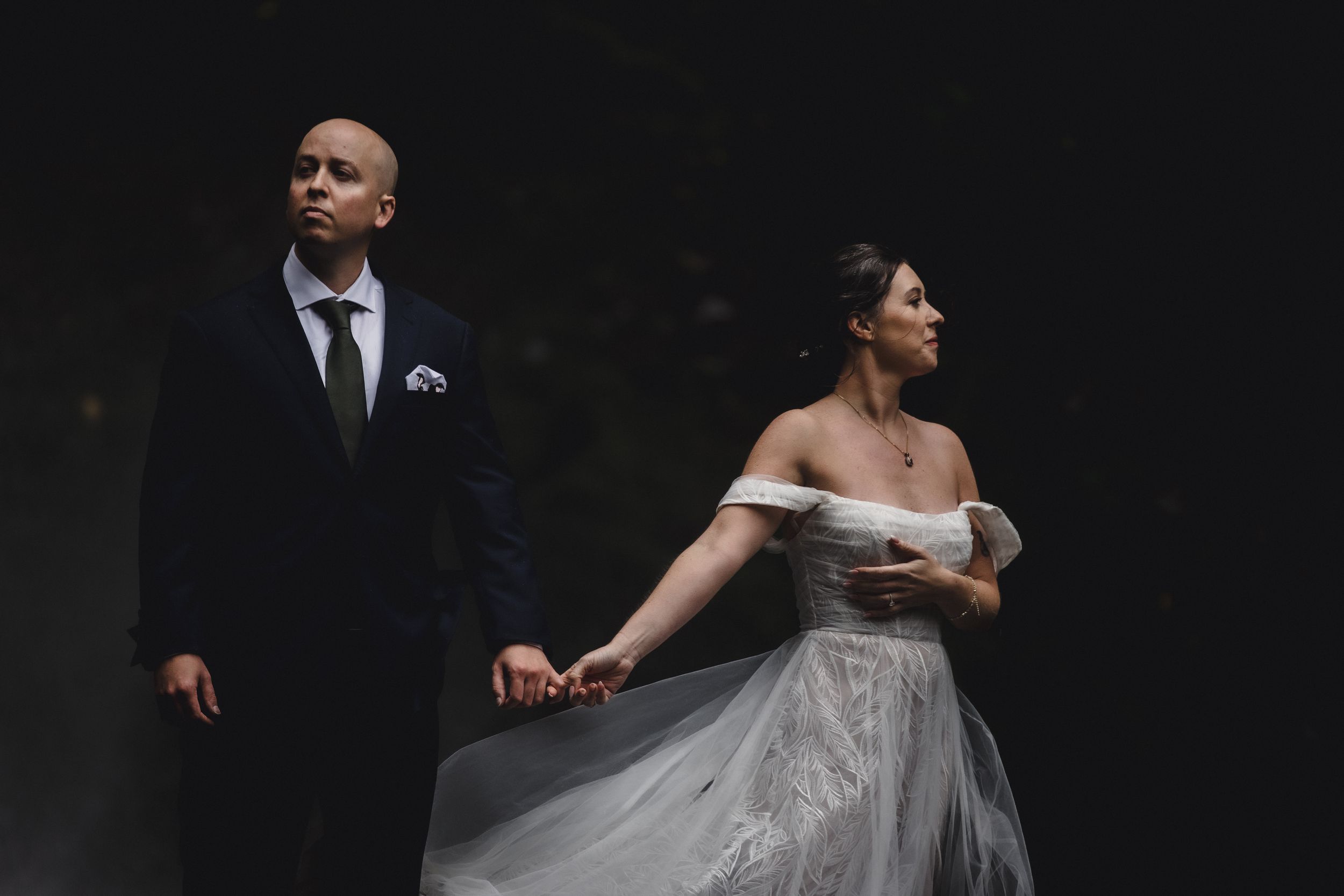 A couple in wedding attire stands close together in a dark forest setting.