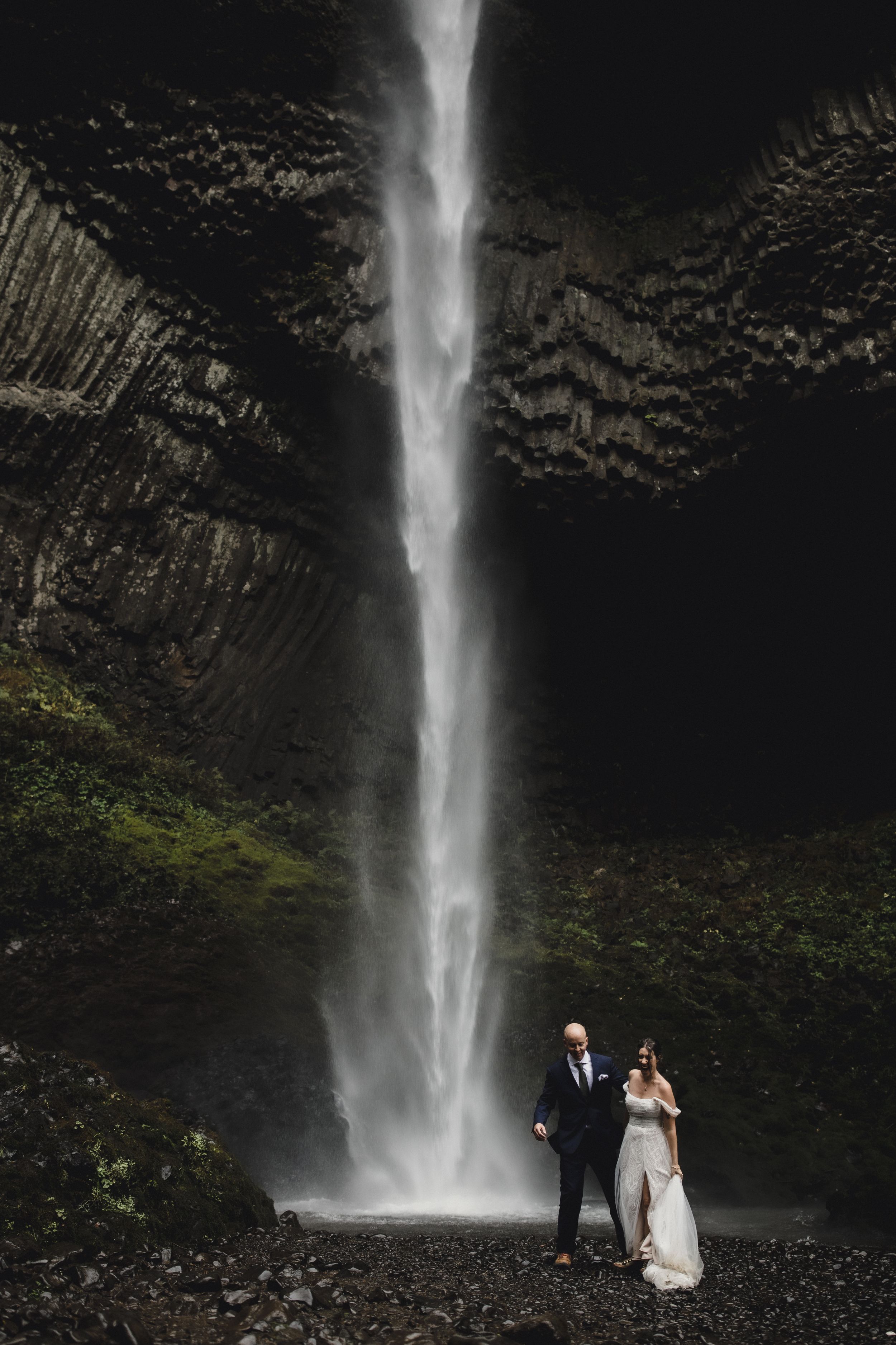 A dramatic wedding portrait with a towering waterfall cascading down dark cliffs in the background.