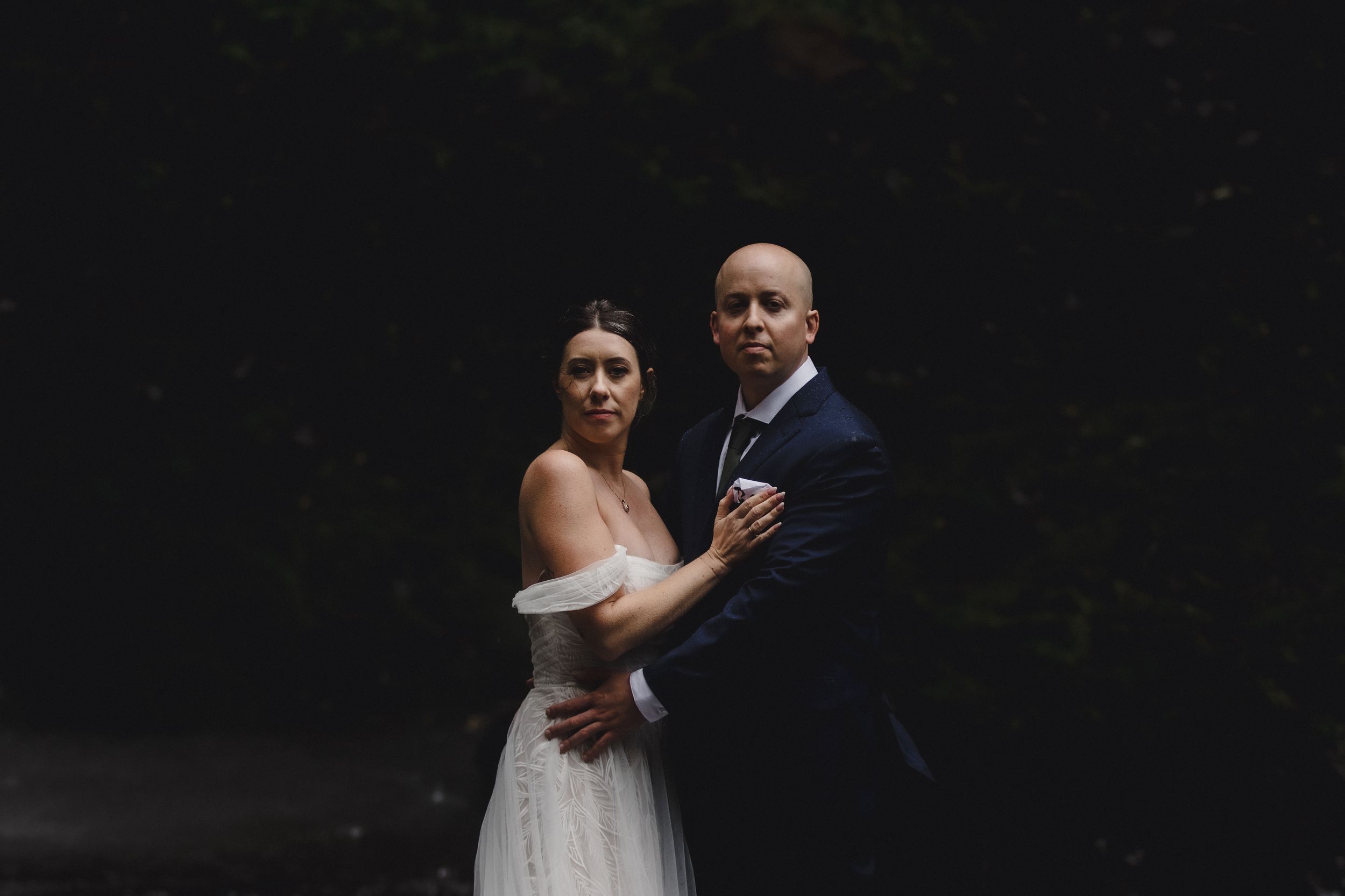 Newlyweds share an intimate moment against a dramatic waterfall backdrop in a forest setting.