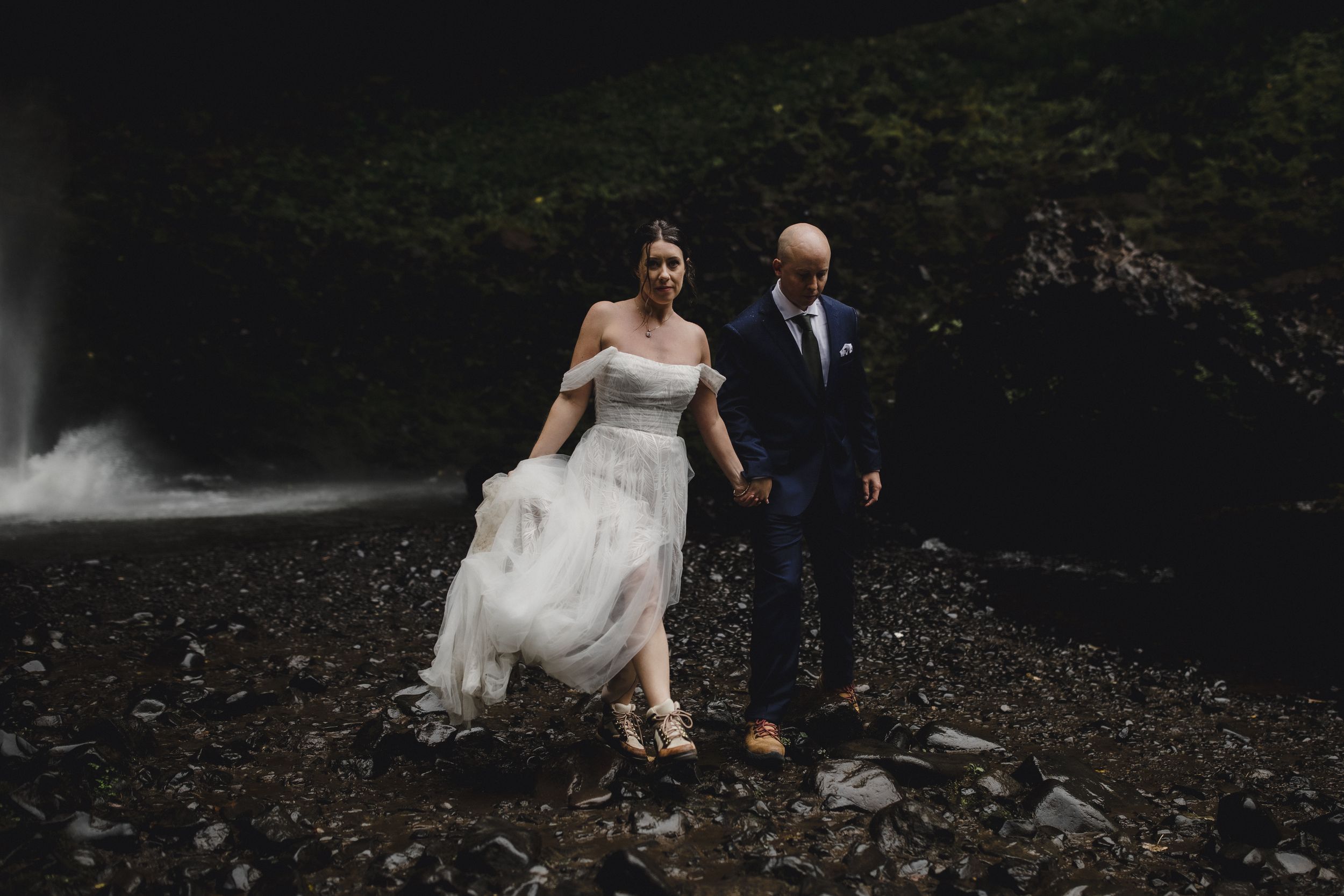Wedding couple stands on rocky terrain with a cascading waterfall creating a dramatic backdrop in the forest.