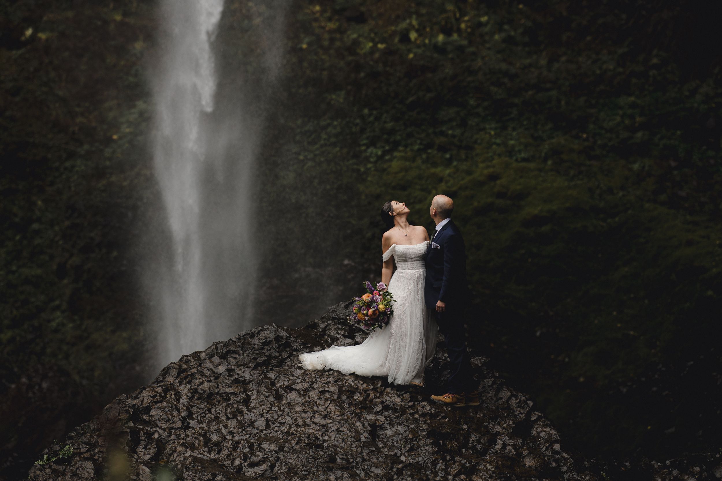 A couple poses near a majestic waterfall in a lush forest setting during what appears to be a wedding photoshoot.