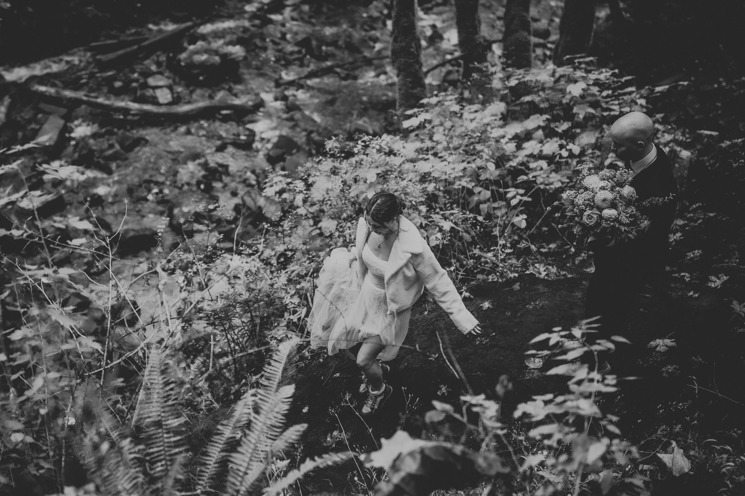 A black and white photograph of a couple embracing in a lush garden setting with natural lighting.