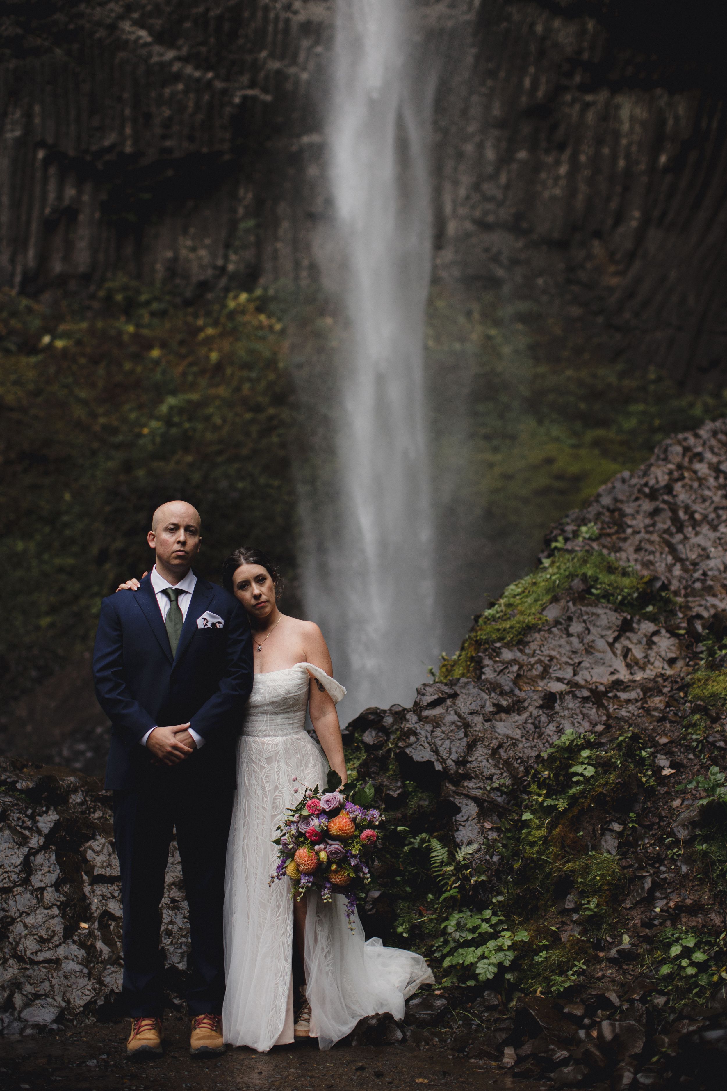 A couple poses for wedding photos in front of a majestic waterfall surrounded by rocky cliffs and lush greenery.