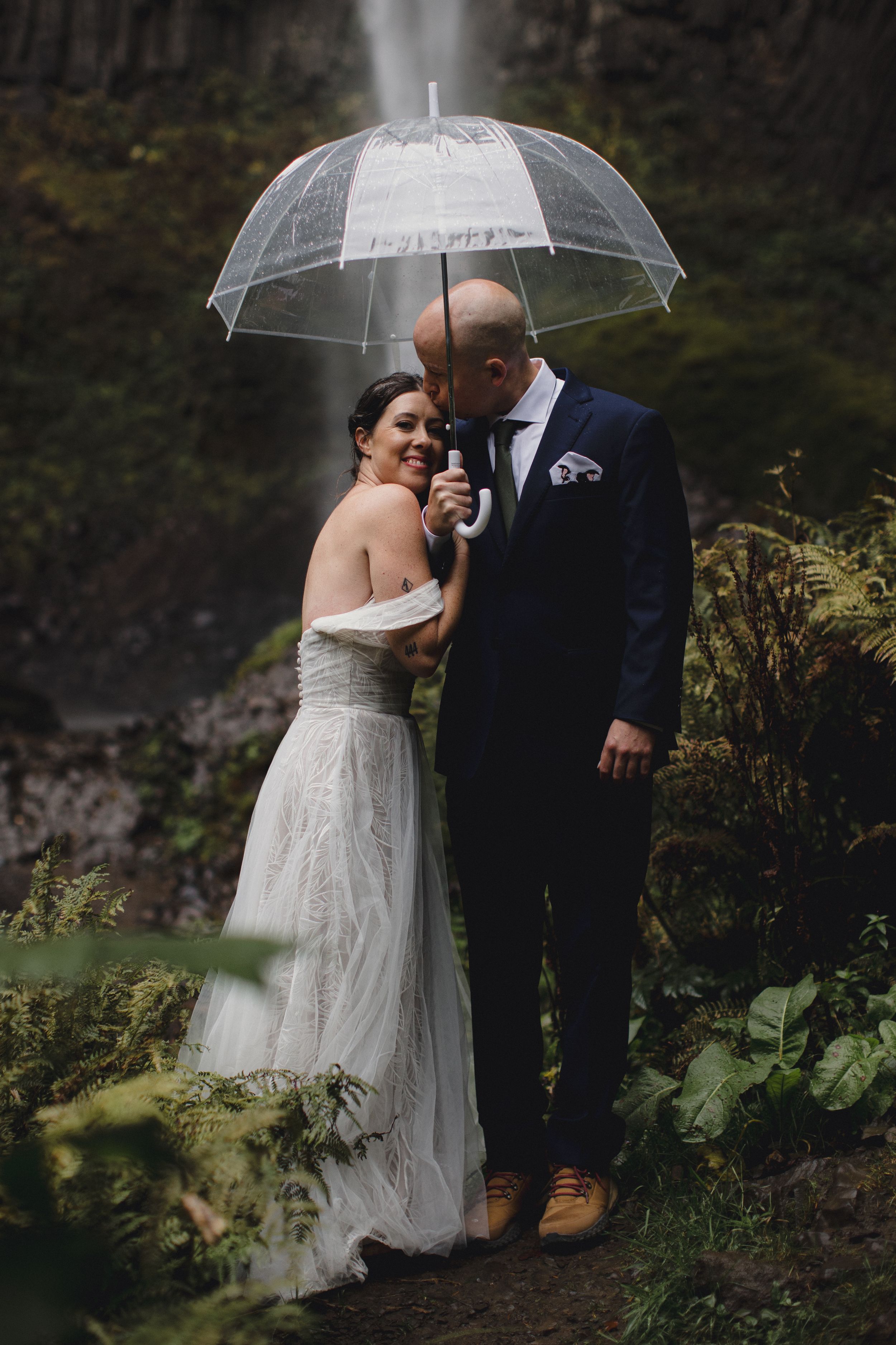 A romantic wedding couple shares an intimate moment under a clear umbrella in a lush garden setting.