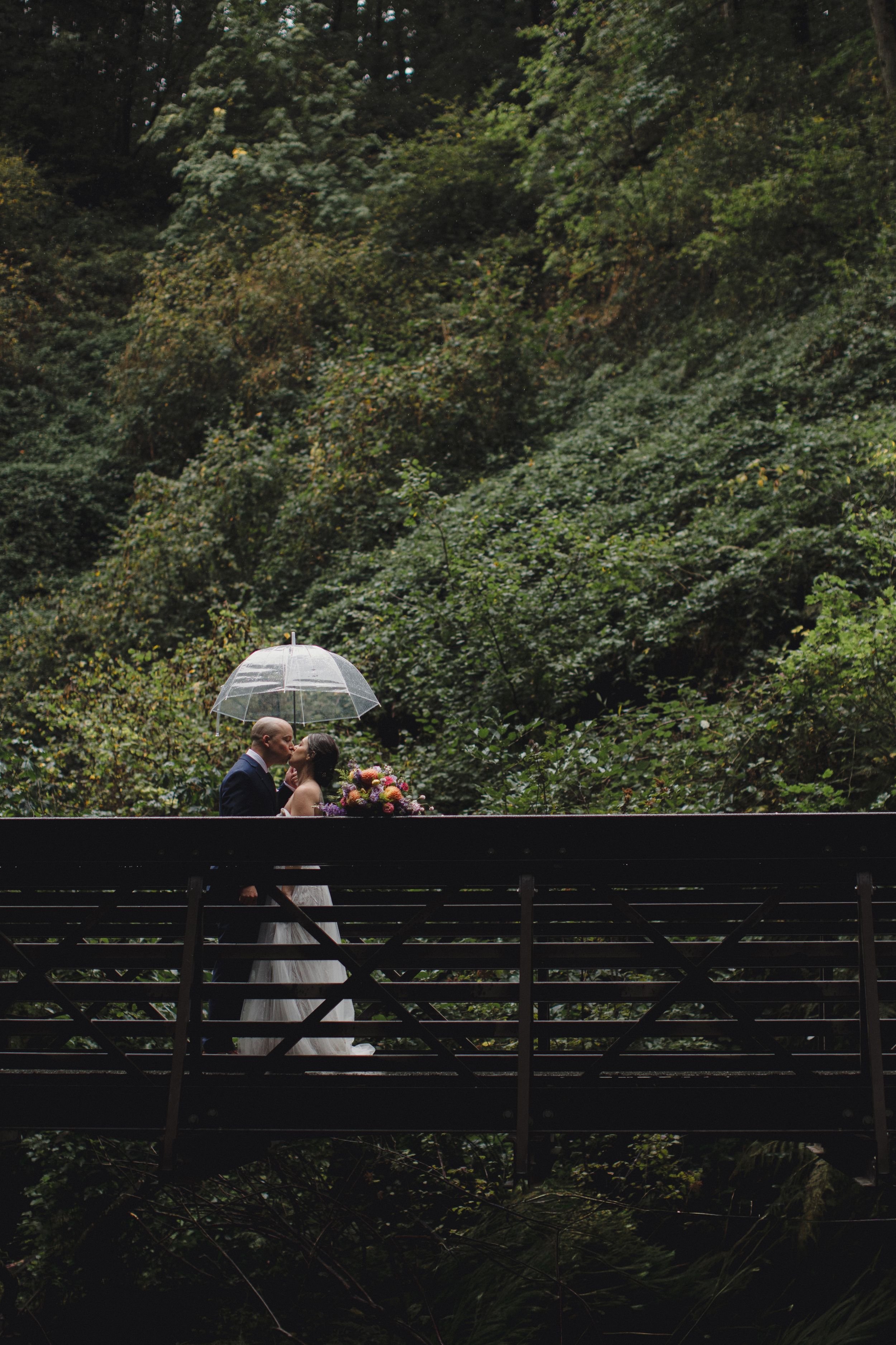 A couple shares an intimate moment under an umbrella on a wooden bridge surrounded by lush greenery.