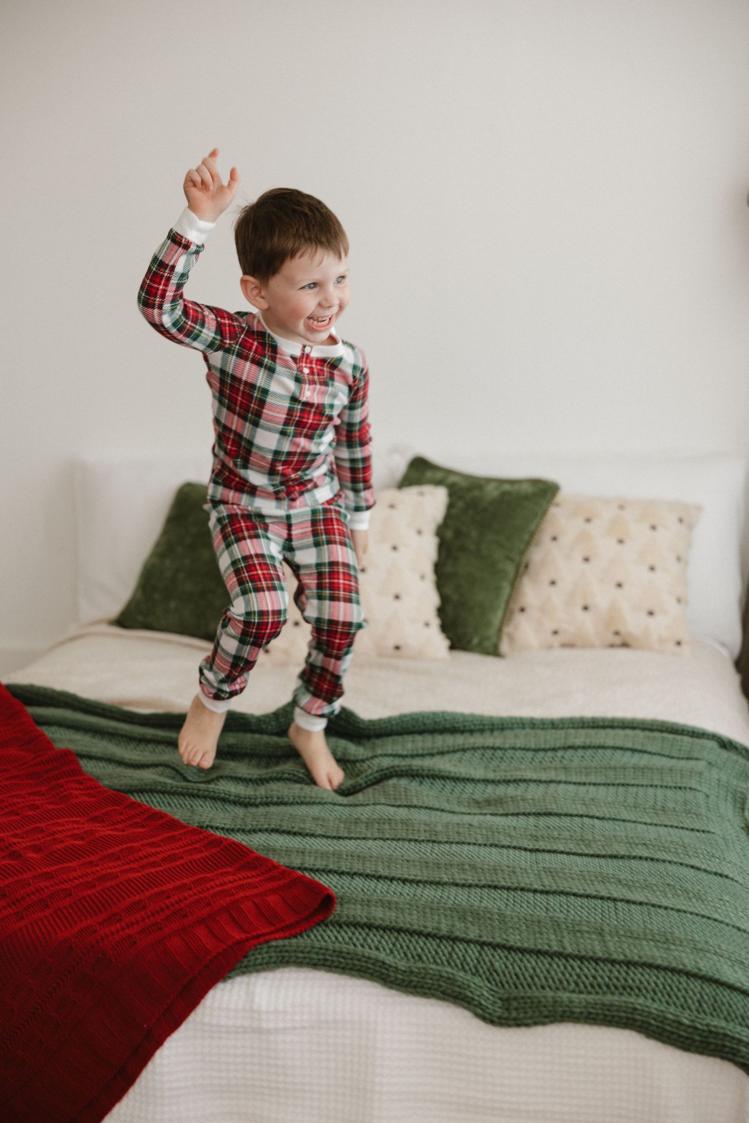 A child in plaid pajamas jumps playfully on a bed with green and red blankets and decorative pillows.