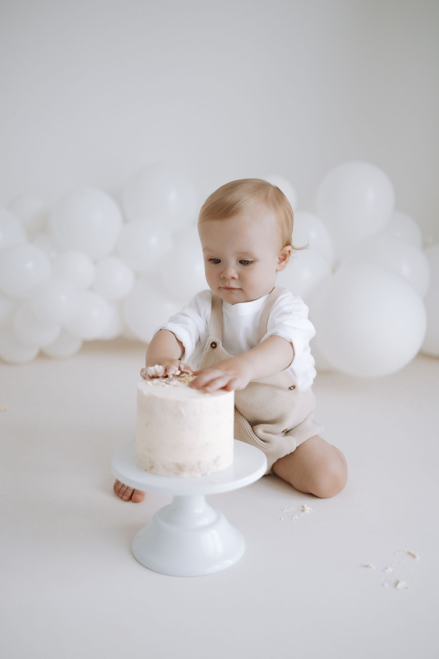 White minimalist cake smash photo session with balloons and a small white cake on a light blue stand.