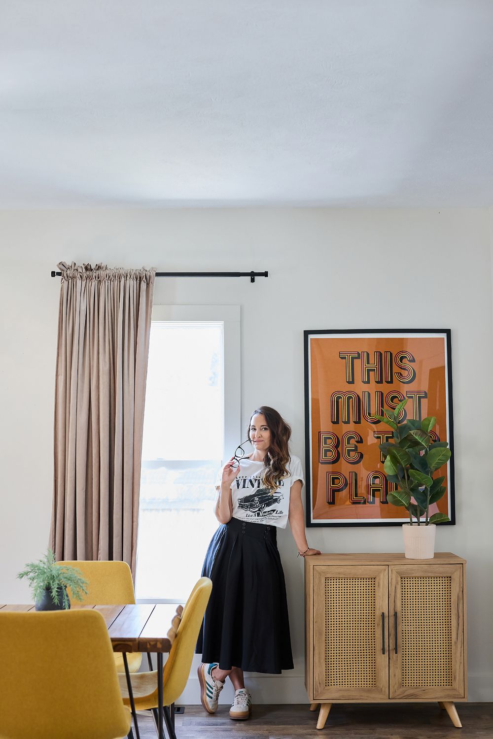 Woman in black skirt and white tee leans against a rattan cabinet beside a bold orange motivational art print.
