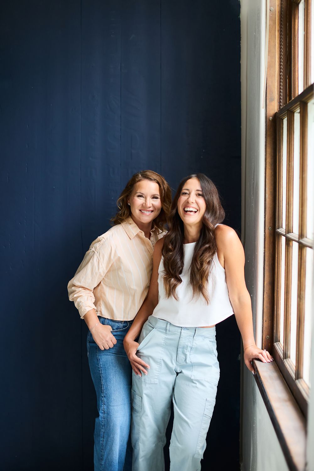 Two people laughing and posing against a navy blue wall with windows.