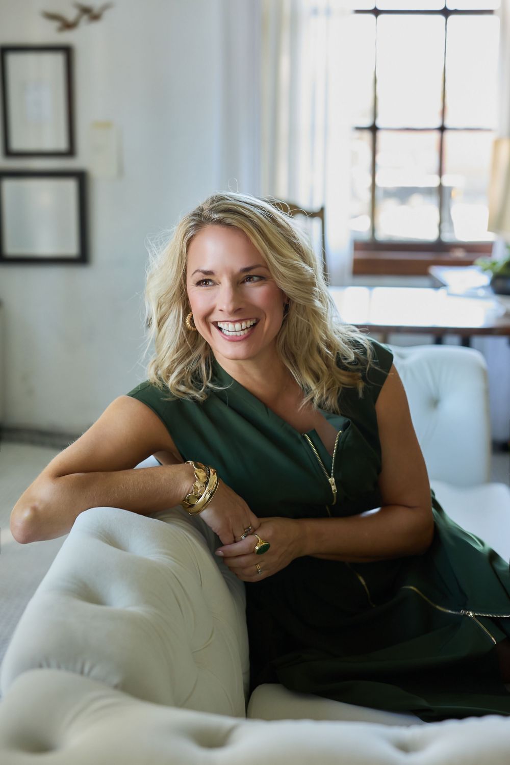 Smiling woman with wavy blonde hair in a green dress relaxes on a tufted white sofa in a bright room with large windows.