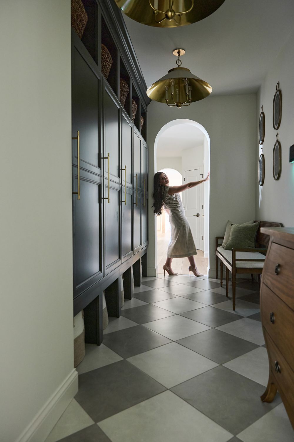 Woman in white dress dancing in elegant hallway with checkered floors, dark cabinetry, and brass pendant lights.
