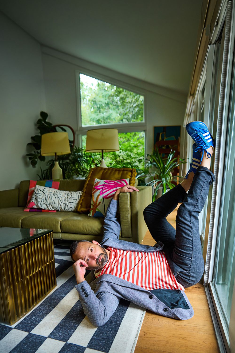 Man lying on striped pillow on checkered rug, legs raised, wearing blue sneakers, in a cozy living room with green sofa.