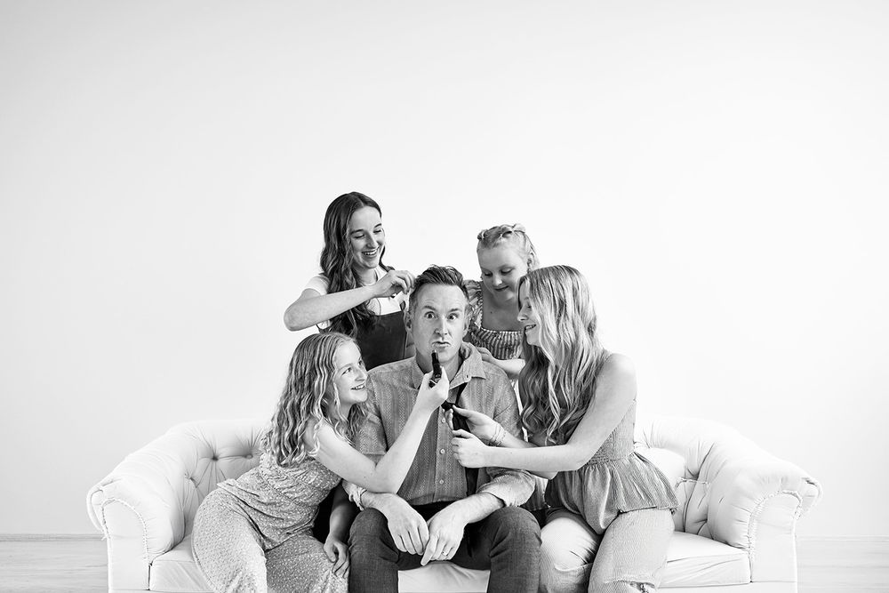 Black and white photo of a family of five laughing together on a white couch in a bright studio setting.