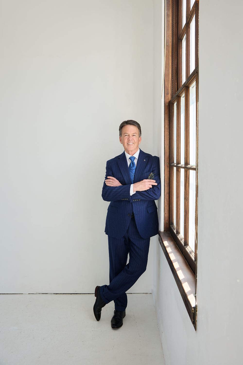 Man in navy pinstripe suit with arms crossed, leaning against window in bright white room.