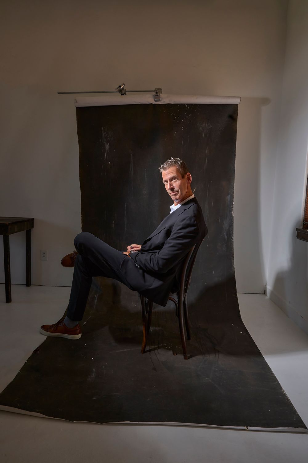 Middle-aged man in dark suit seated on chair against dark backdrop in professional studio portrait setting.