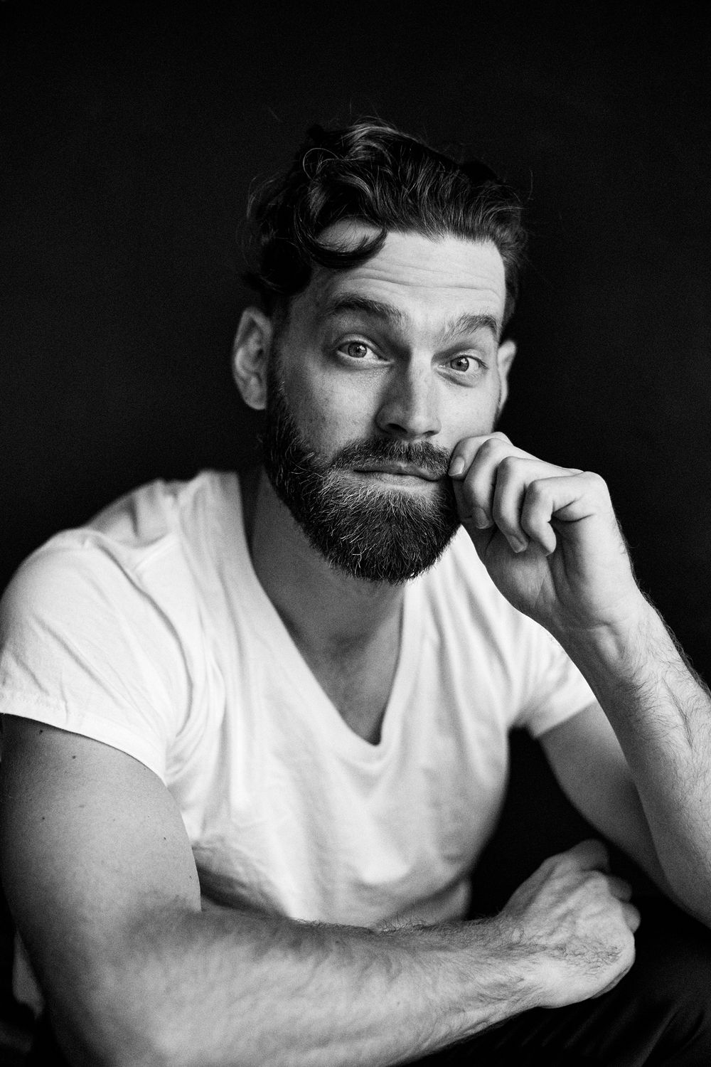 Portrait of a bearded man in a white t-shirt making a playful expression against a dark background.