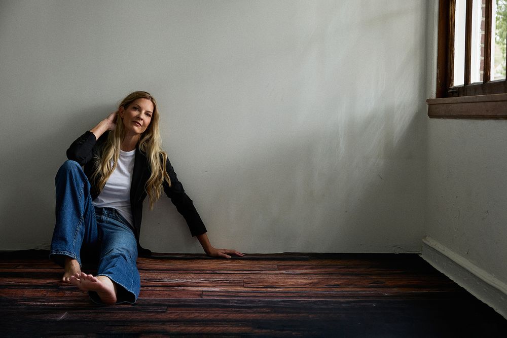 Young woman with long blonde hair sitting on dark hardwood floor, smiling, wearing jeans and black jacket.