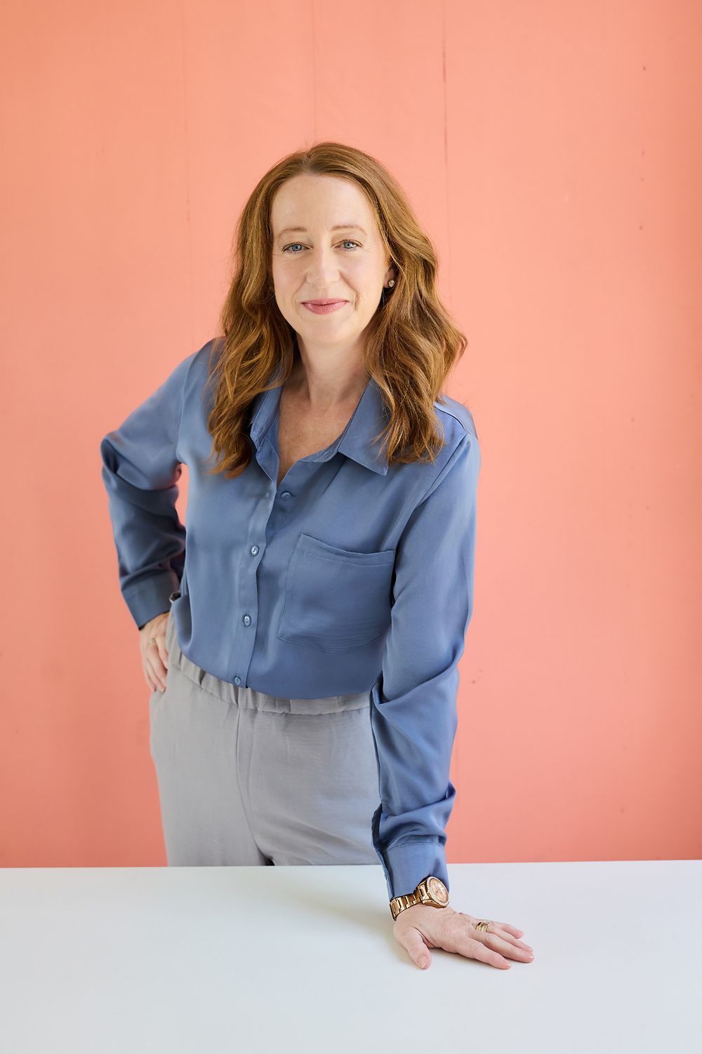 Professional woman in blue silk blouse and gray pants posing confidently against a coral pink background.