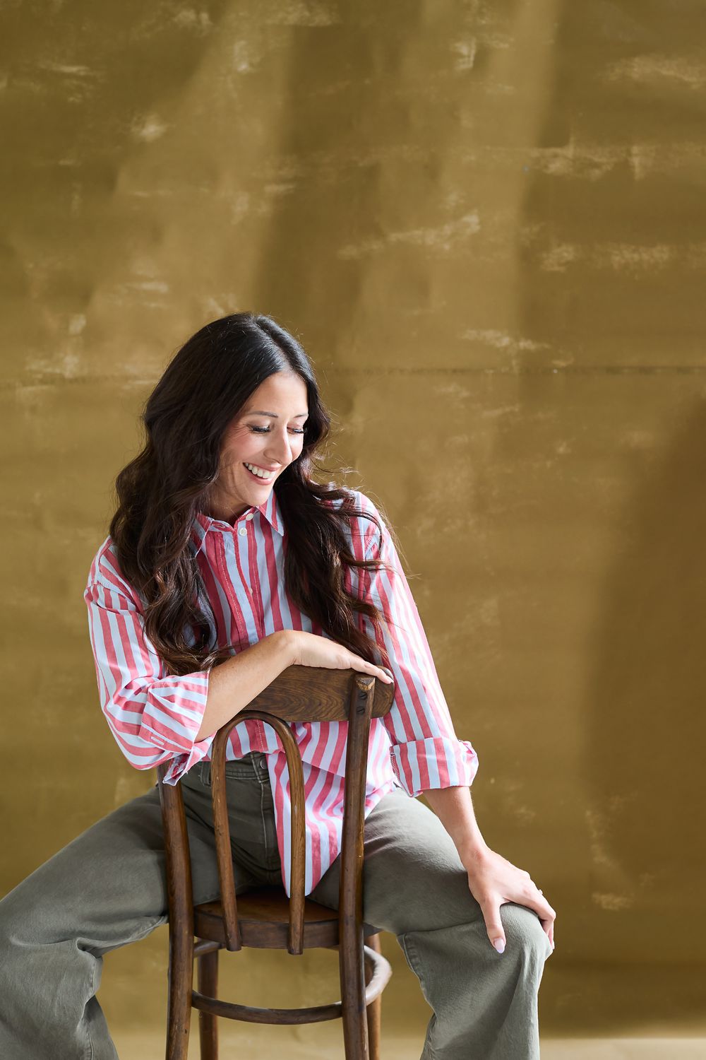 Woman in striped shirt posing artistically with wooden chair against gold background.