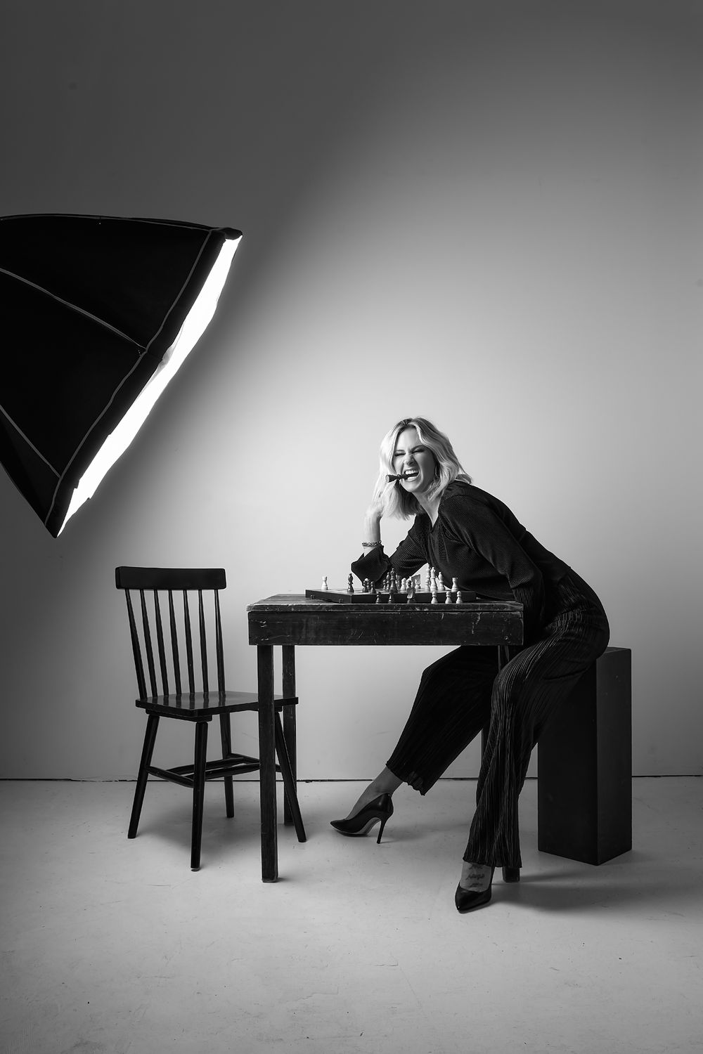 Black and white studio photo of a smiling blonde woman in black outfit sitting at a chess table with a softbox light.