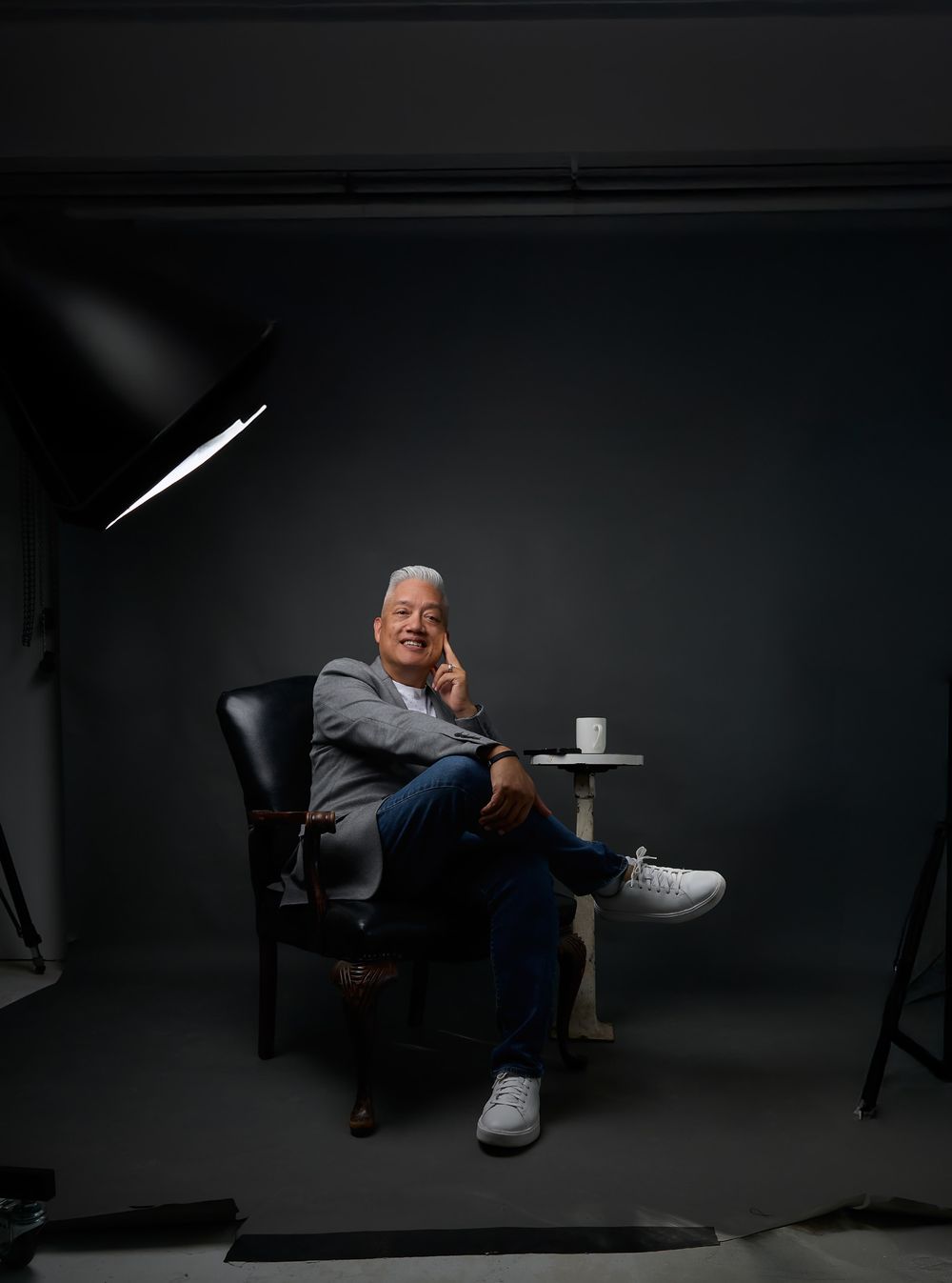Professional studio portrait of person in grey jacket seated in dark setting with dramatic lighting.