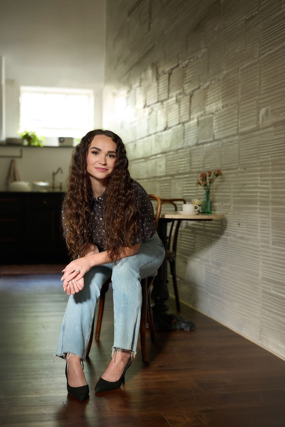 Young woman with curly hair sits on a chair by a brick wall, wearing jeans and heels in a cozy interior.