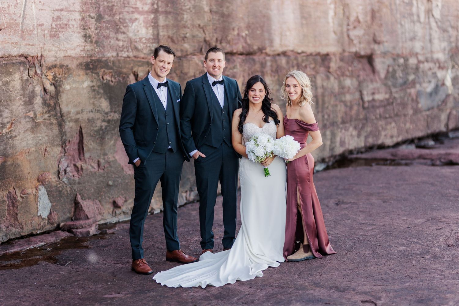 Wedding party poses against rustic stone wall in formal attire with bridesmaids in dusty rose dresses.