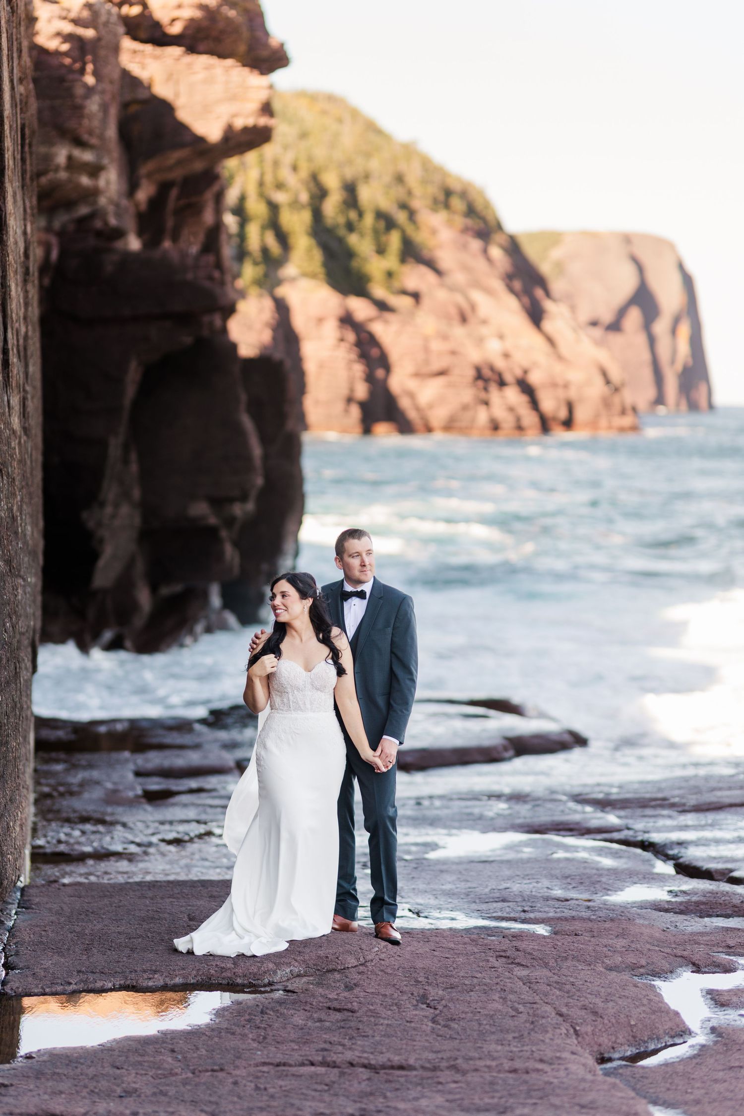 A couple poses for wedding photos on rocky cliffs overlooking the ocean at sunset.