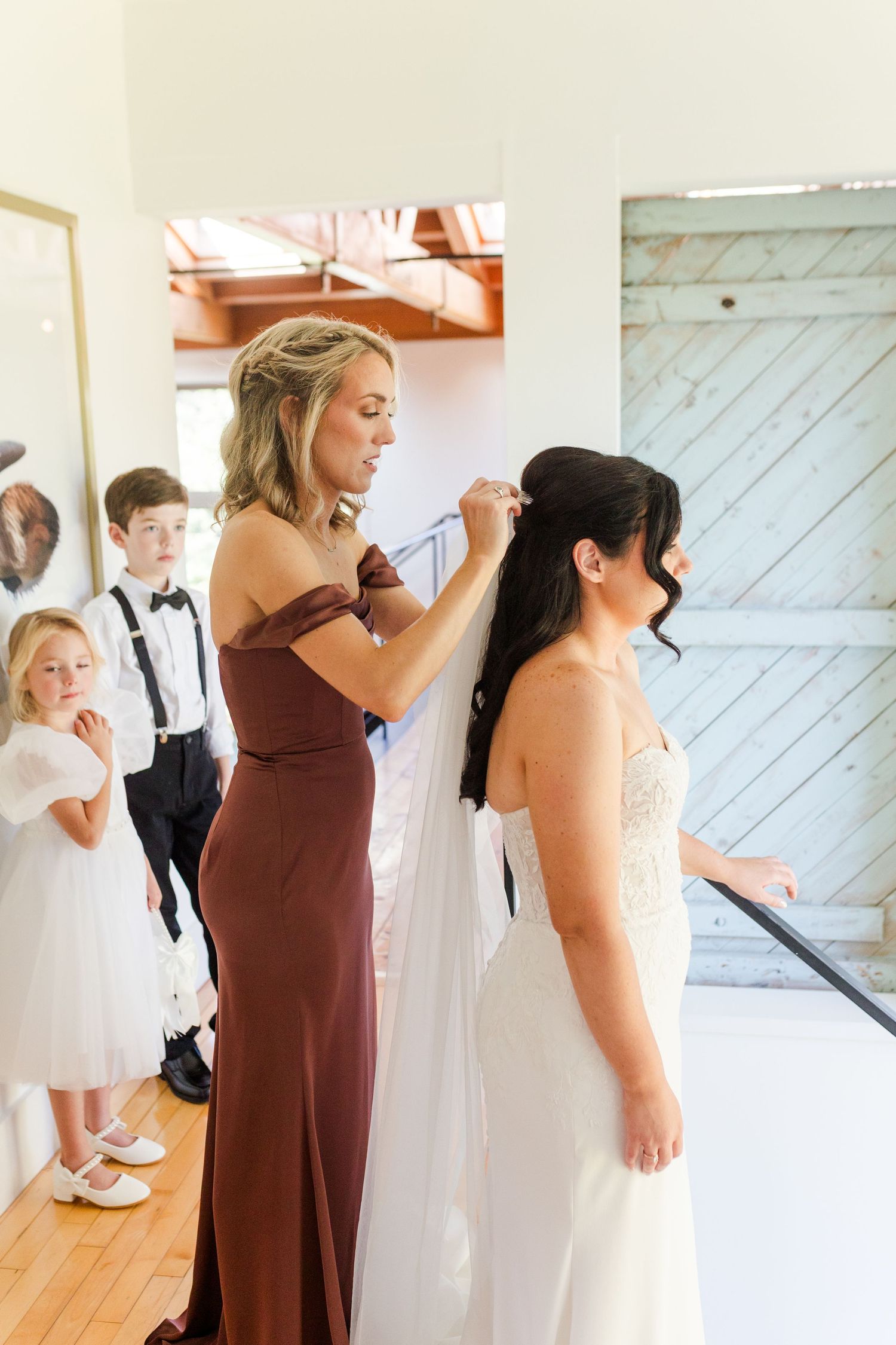 Wedding party members help with final dress preparations in a bright white room.