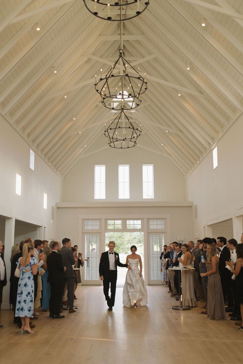 Wedding guests line the walls of an elegant white chapel with vaulted ceilings and chandeliers.