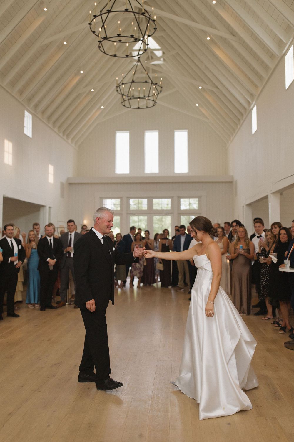 Couple dancing in an elegant white reception hall with high vaulted ceilings and gathered guests watching.