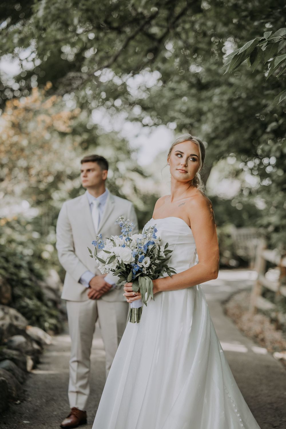 A wedding couple stands on a tree-lined path with the bride in white holding a blue and white floral bouquet.