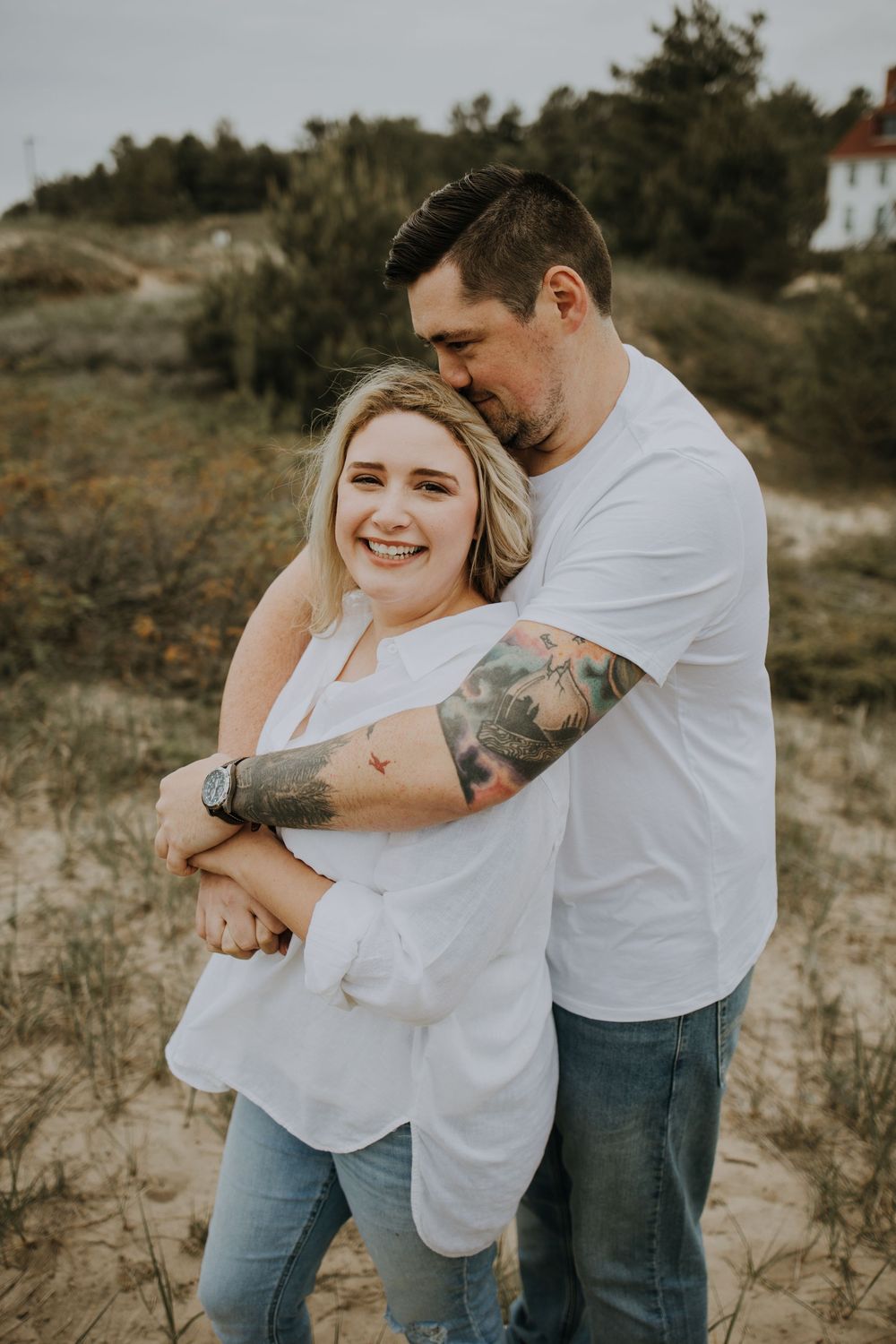 A couple in white shirts and jeans embrace playfully in a natural outdoor setting with sandy terrain and vegetation.