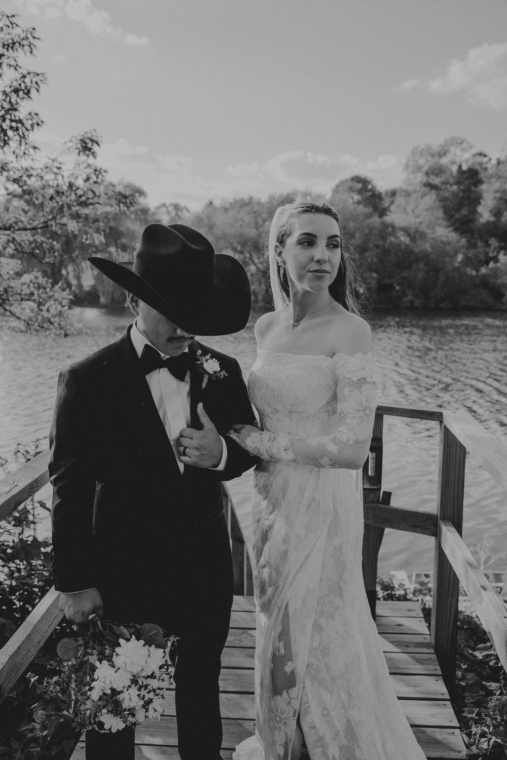Black and white wedding photo of bride in lace gown and groom in tuxedo with cowboy hat on wooden dock by lake.
