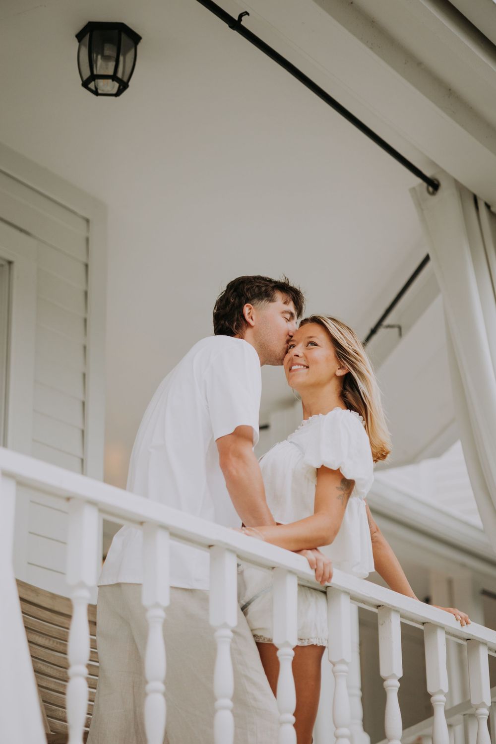 Couple in white outfits sharing a romantic moment on a white balcony with railing.