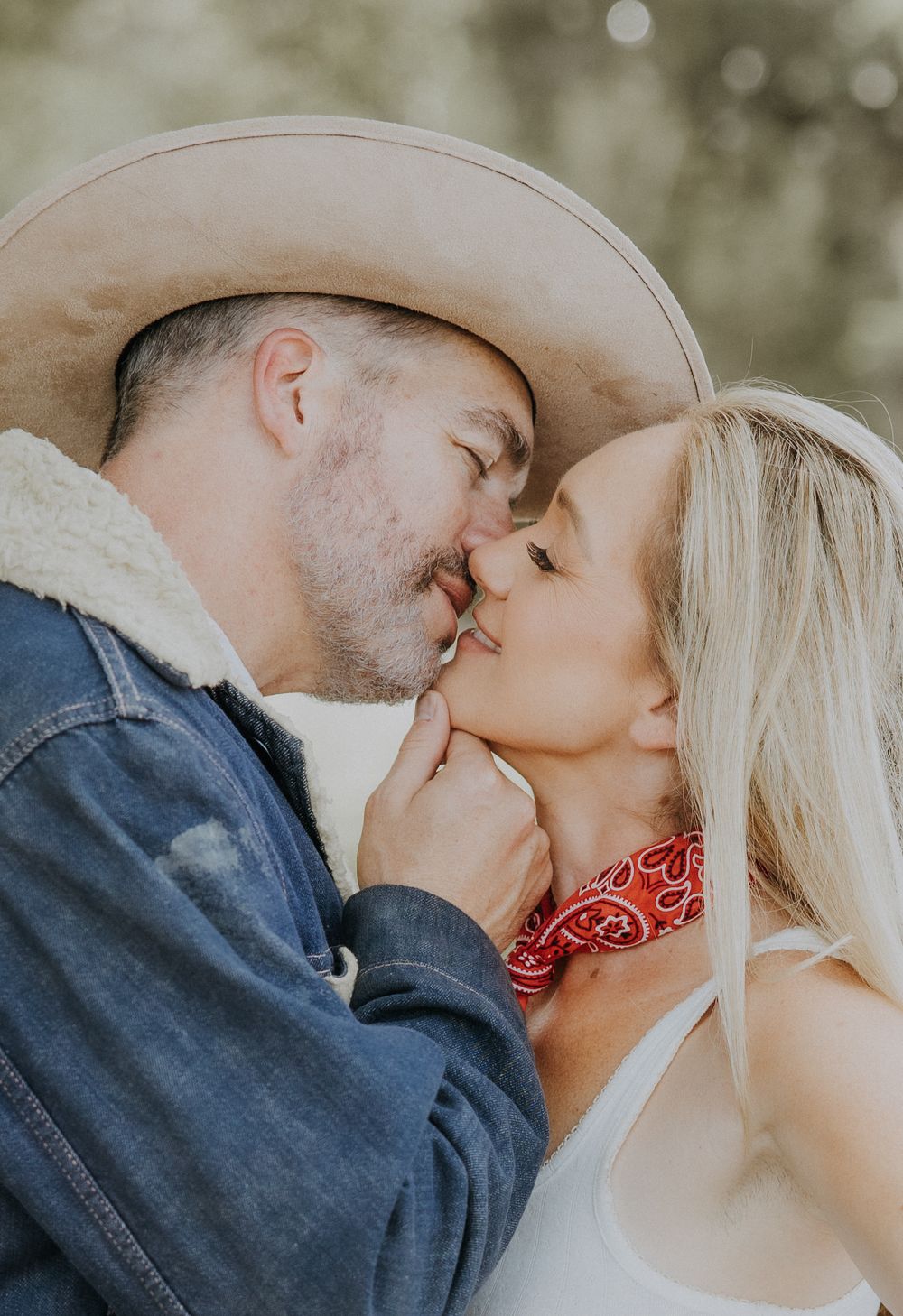 A couple shares a romantic kiss outdoors, man wearing a cowboy hat and denim jacket, woman in white top with red bandana.